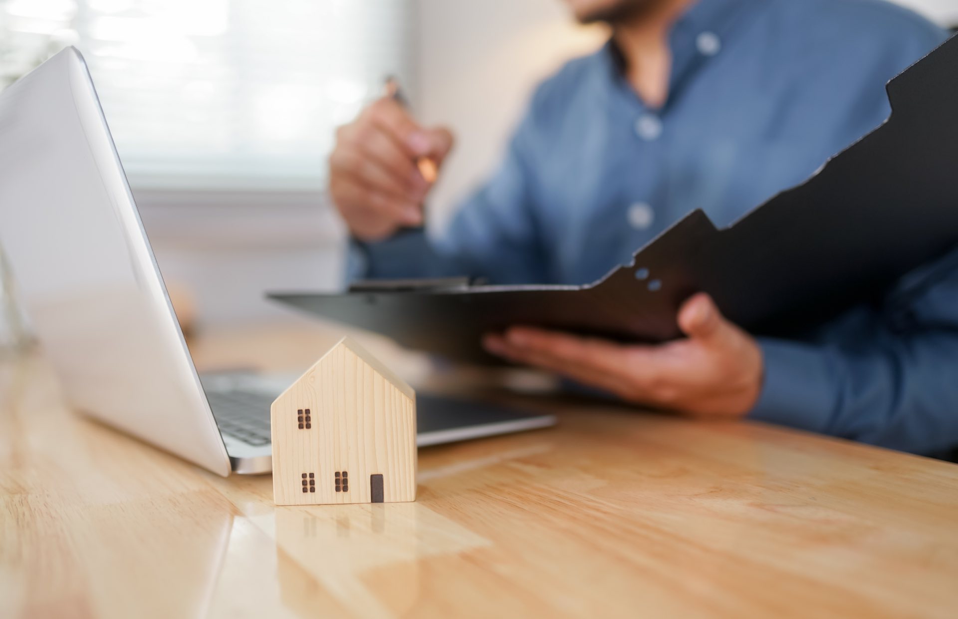 Person holding a pen and a folder with a laptop in front of him and a small wooden house