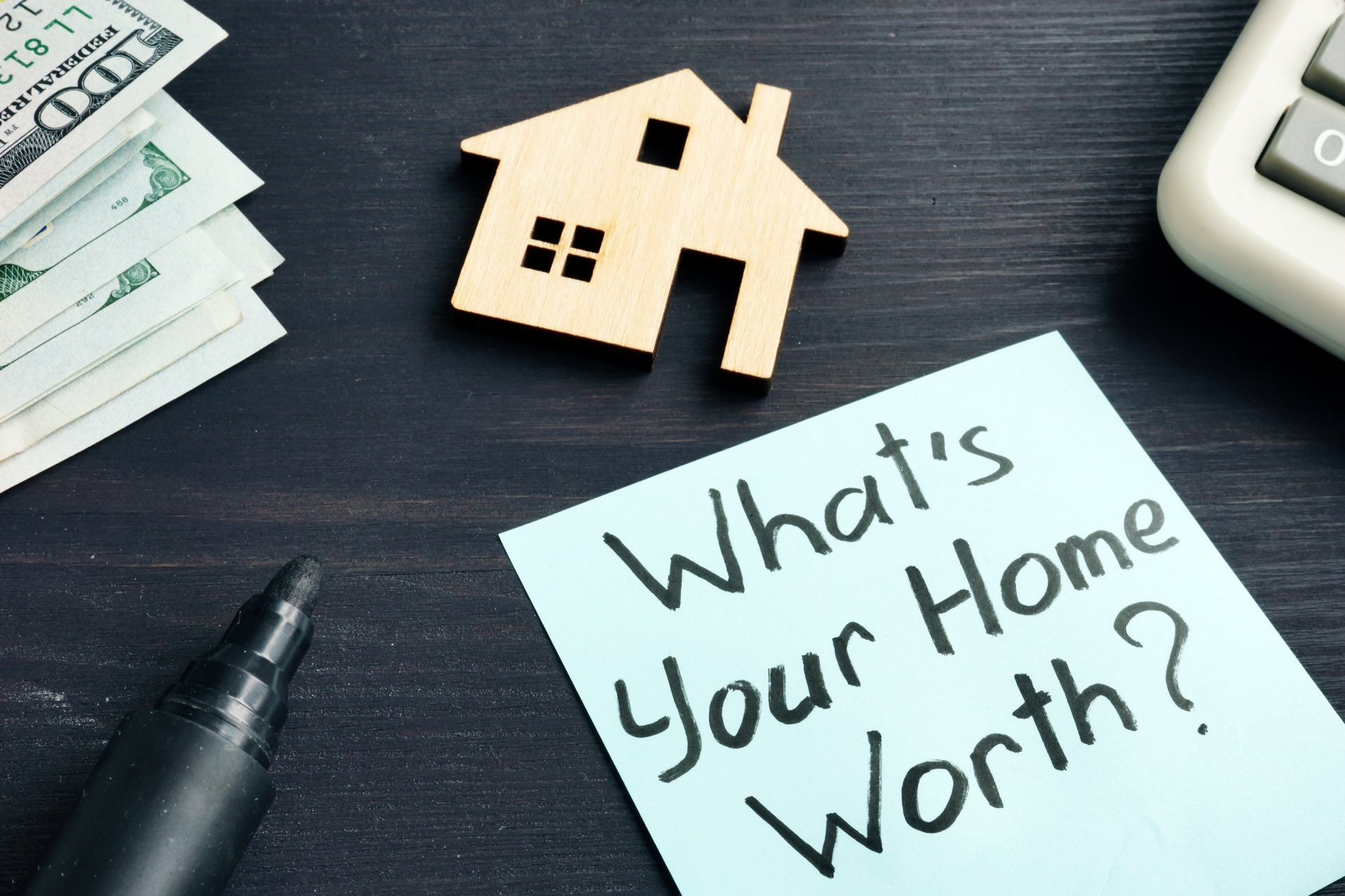 What's your home worth? written on a sticky note next to money, a small wooden house, a pen and a calculator
