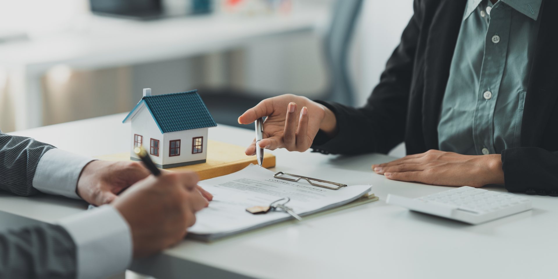 Close up of two people sitting at a desk holding pens in front of paperwork and a small wooden house